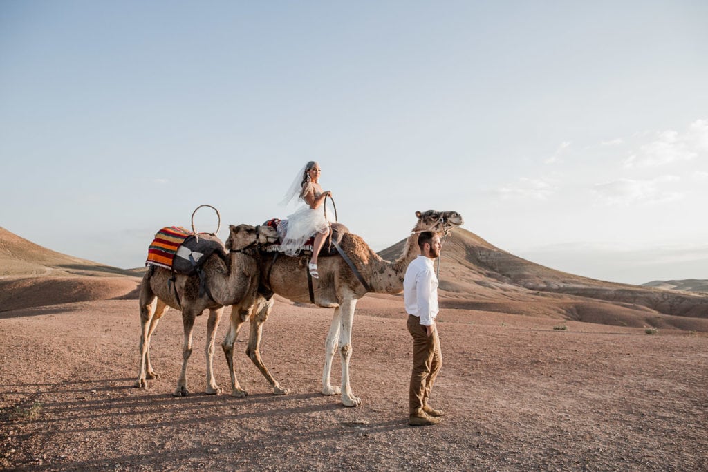 Bride sits atop camel while groom stands next to her during Morocco destination desert wedding couple portraits