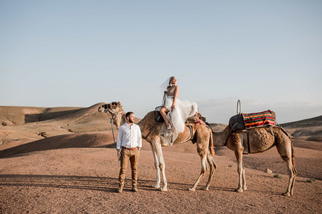 Bride sits atop camel while groom stands next to her after Morocco Marrakech wedding.