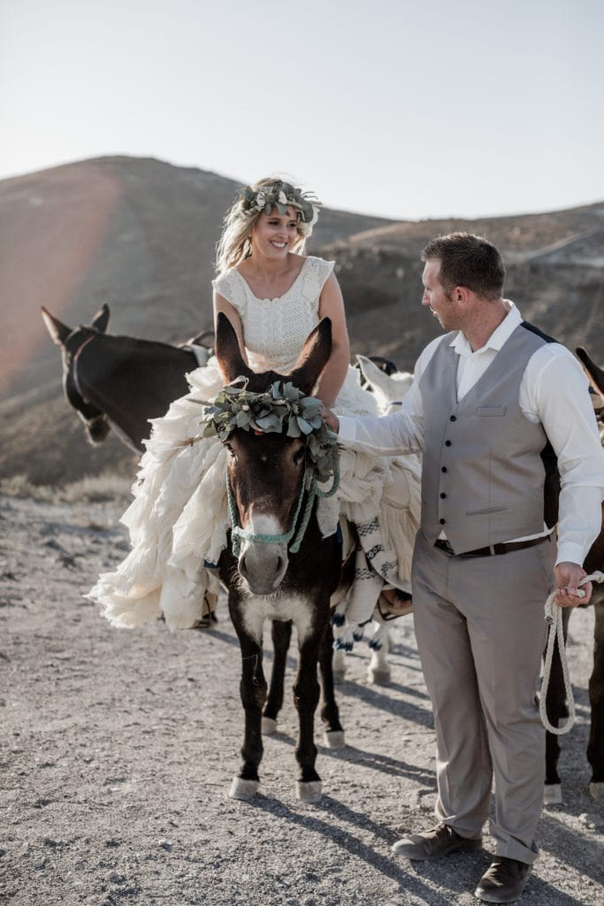 Bride sits atop mule while groom stands next to her in Greece cliff sides