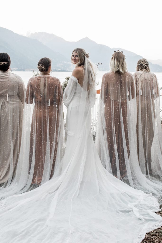 Bride looks over shoulder as she stands with bridesmaids, all wearing pearl-netted capes