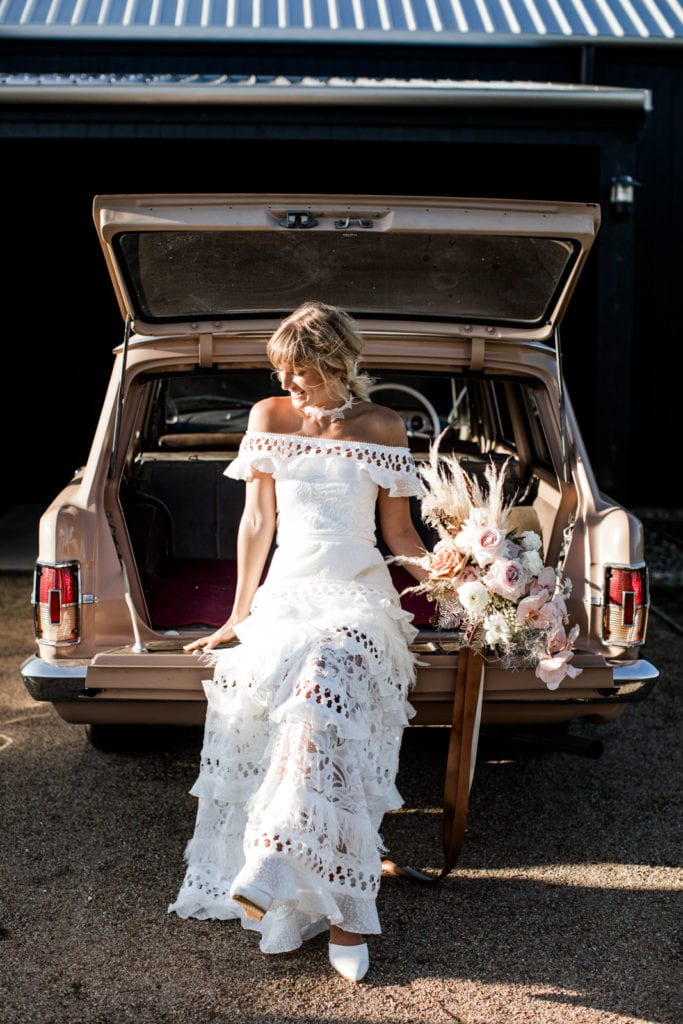 Bride wears bohemian-chic wedding gown while holding her bouquet and sitting on back of vintage car