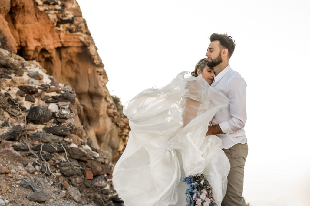 Bride and groom stand together during sunset couples portraits on a cliffside in Santorini