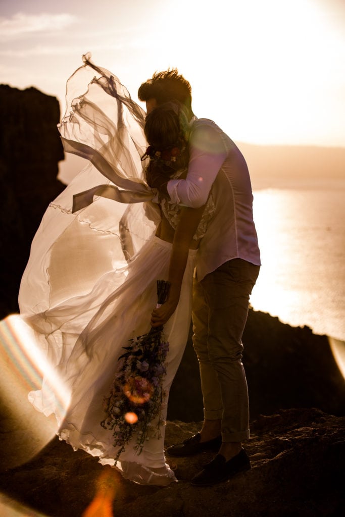 Bride and groom embrace during sunset portraits after wedding ceremony while wind blows bride's dress in Santorini