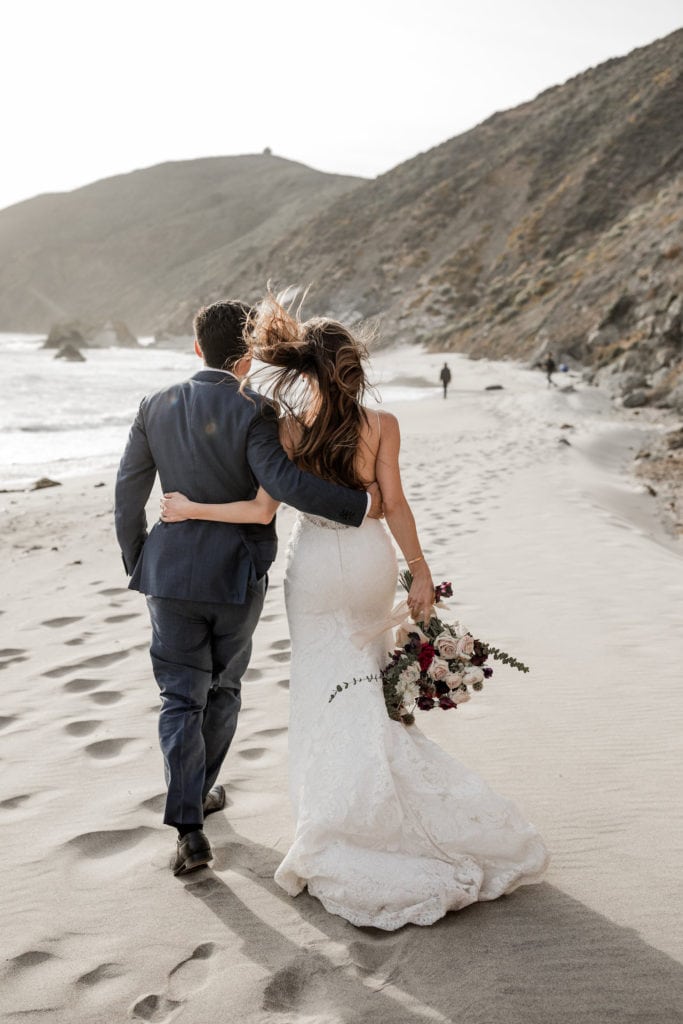 Bride and groom walk down sandy beach in Big Sur, California