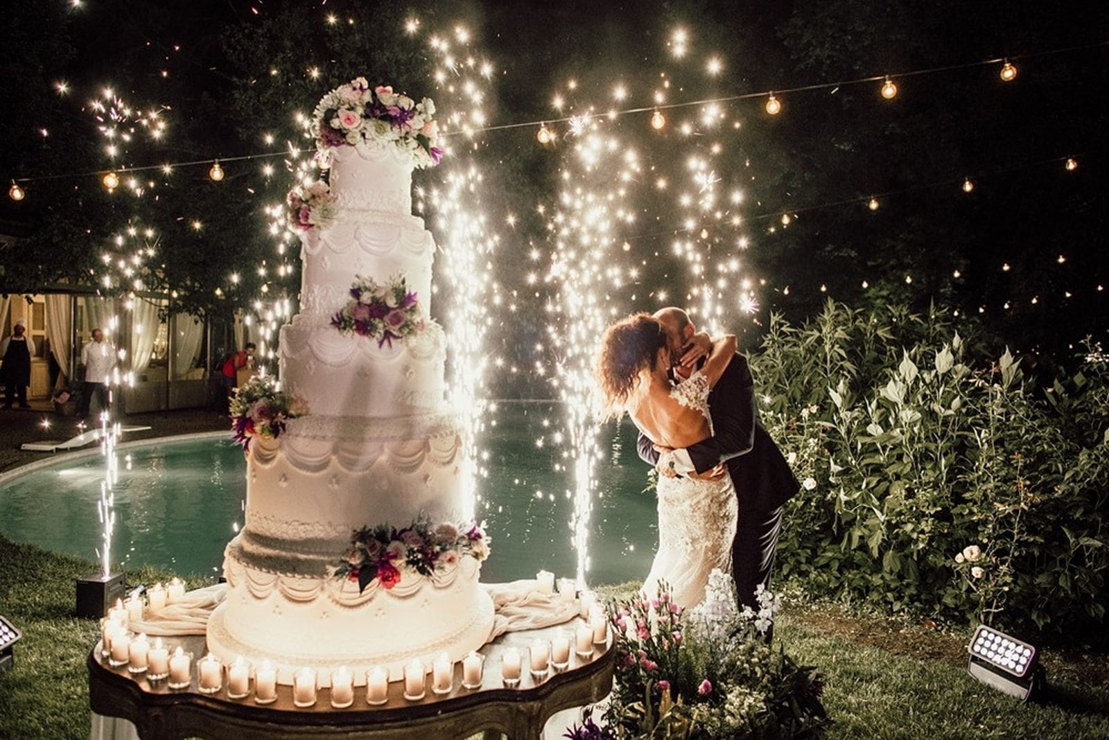 Bride and groom kiss after cutting into wedding cake with fireworks in background