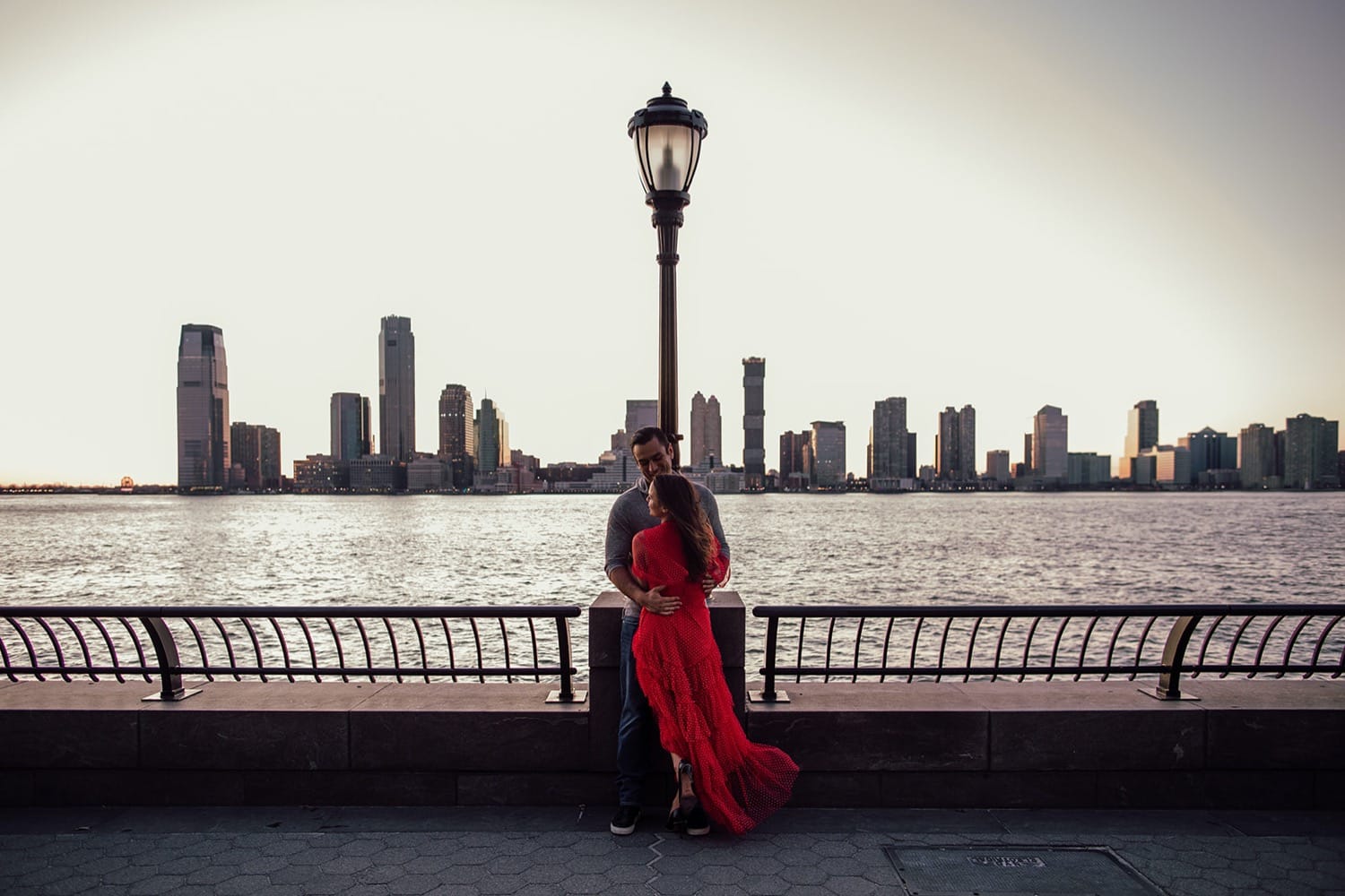 Man and woman hug in front of Hudson River with city skyline in the background during New York City engagement session
