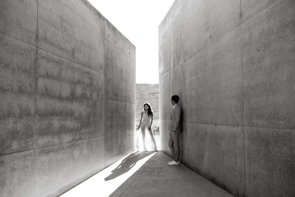 Woman stands between architecture at Amangiri while man looks at her for Amangiri engagement session portraits
