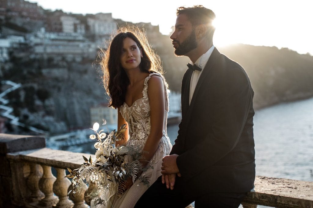 Bride and groom sit on bannister at Villa Astor overlooking Amalfi Coast