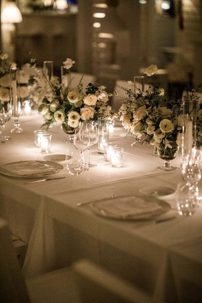 Dinner and glassware line tables with white and cream colored flower centerpieces at a wedding reception. 
