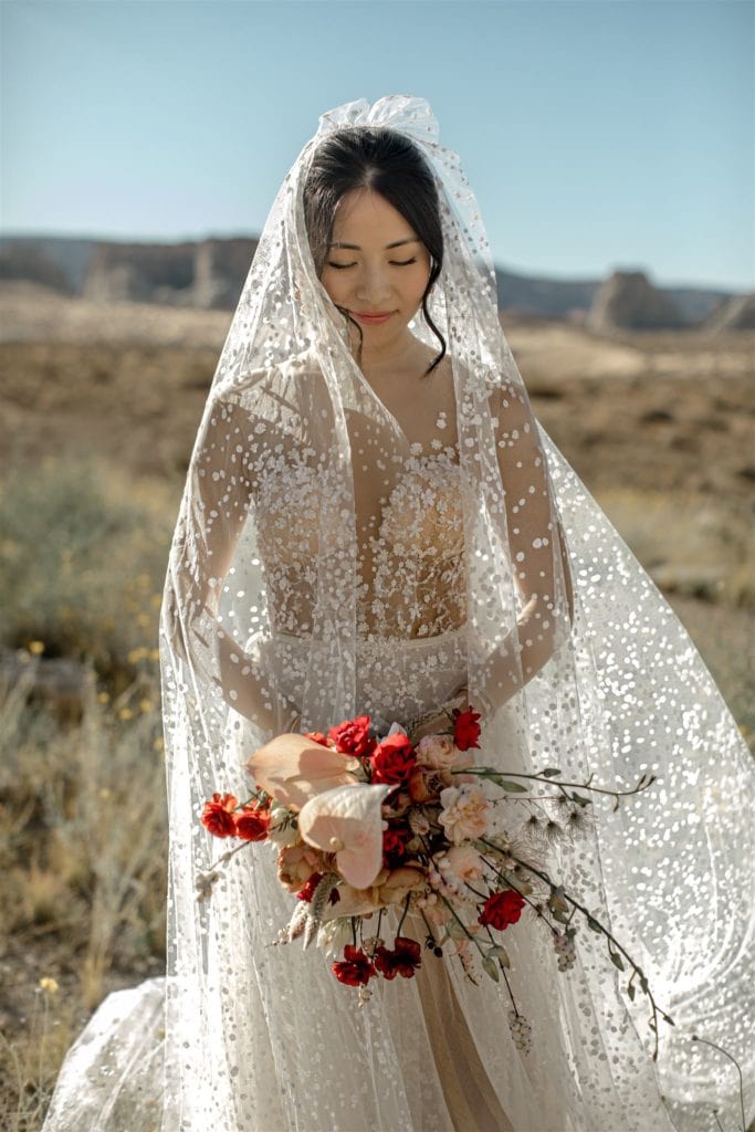 A bride wearing a Berta dress with matching veil looks down at her red and pink bridal bouquet. 