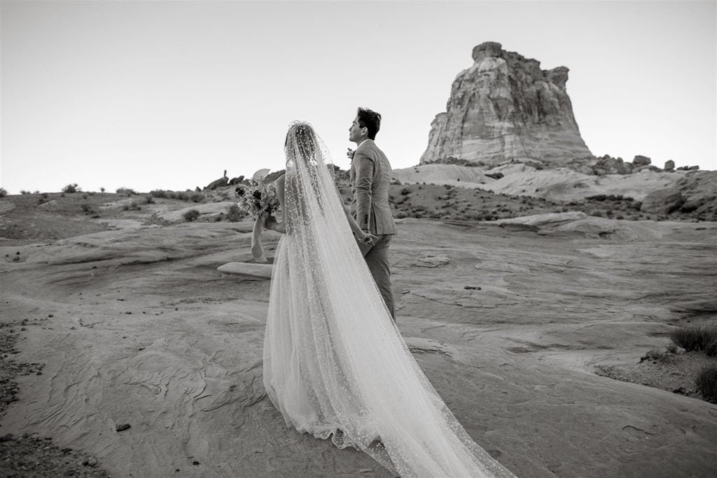 A black and white portrait shows a bride and groom walking through the plateaus of Canyon Point, Utah. 