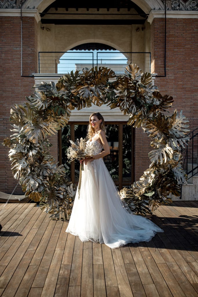 A bride stands in front of a circular arch made of golden palm fronds
