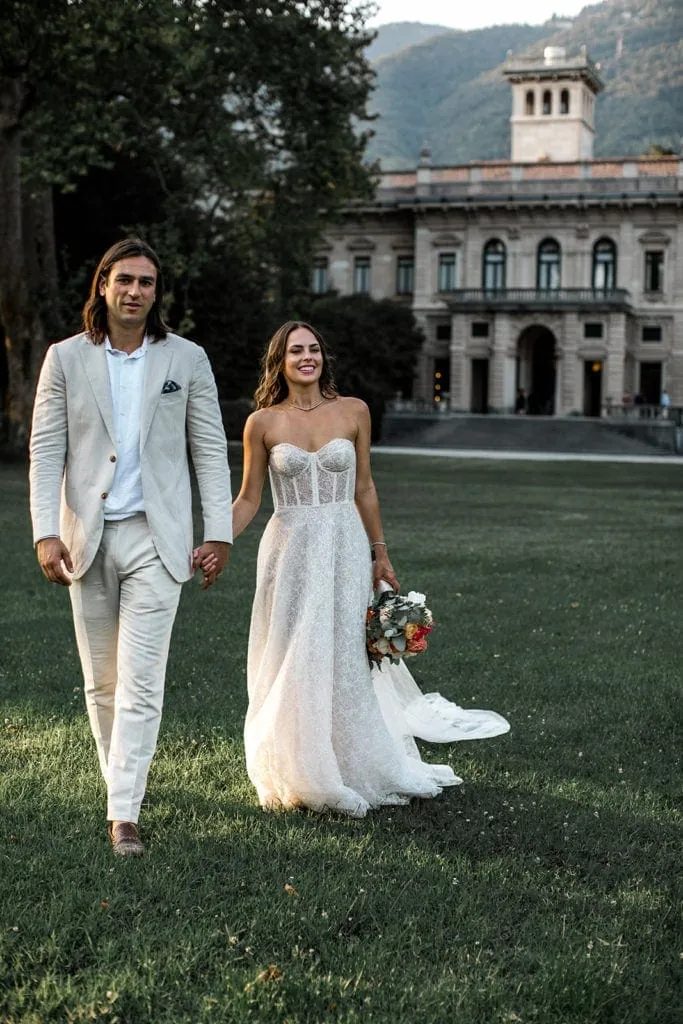Bride and groom, who wears tan suit ideas, walking in field with Villa Erba in background