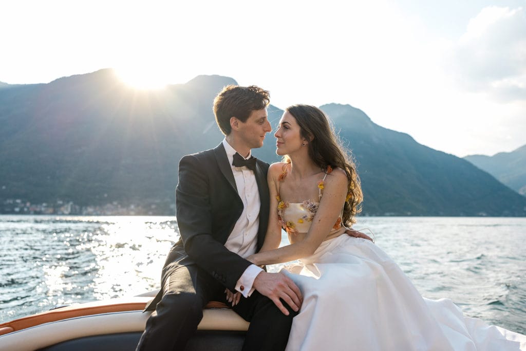 Bride and groom, wearing a black groom's tuxedo, sit on bow of a boat on Lake Como
