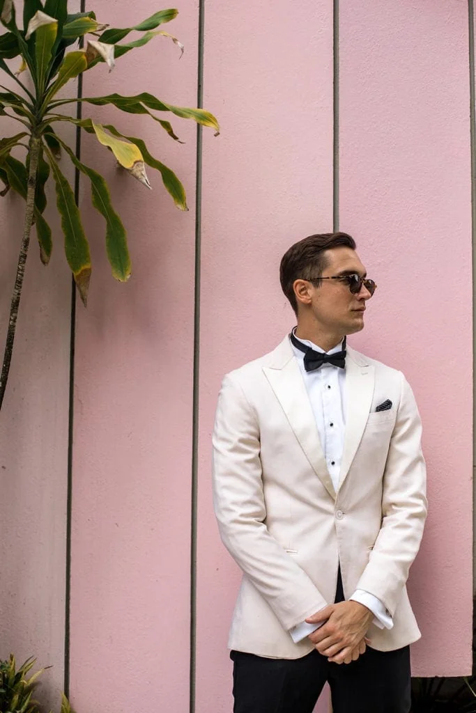 Groom stands in a cream colored tux jacket against a pink wall