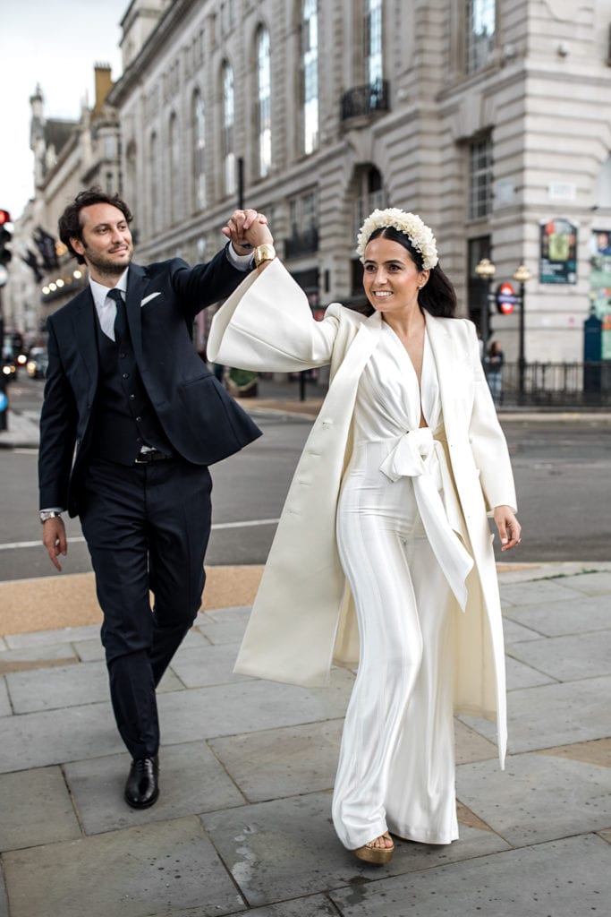Bride in a Galvan jumpsuit spins under groom's arm on streets of London.