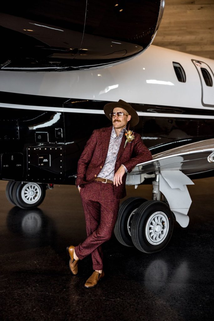 Groom leans against vintage airplane in Daniel George custom suit