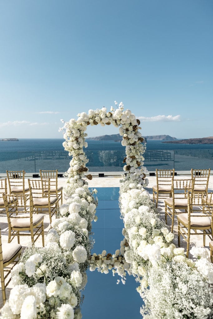 White floral arch and mirrored ceremony aisle in front of waterfront in Santorini Greece
