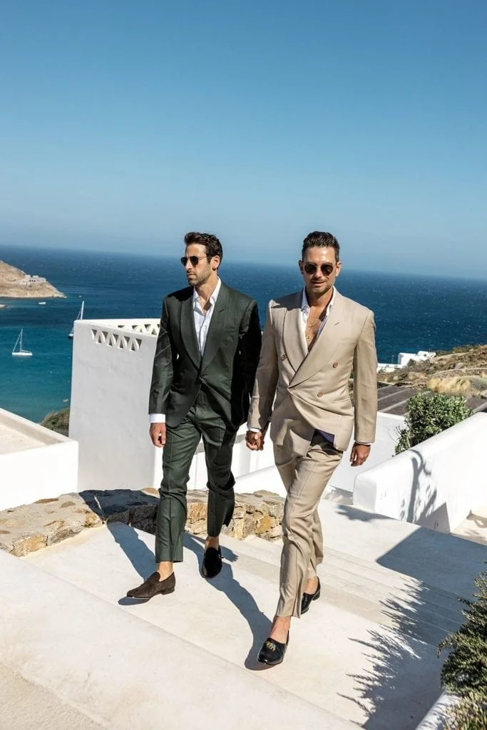 Grooms, both wearing beach wedding clothes for groom, walk together to Mykonos wedding ceremony