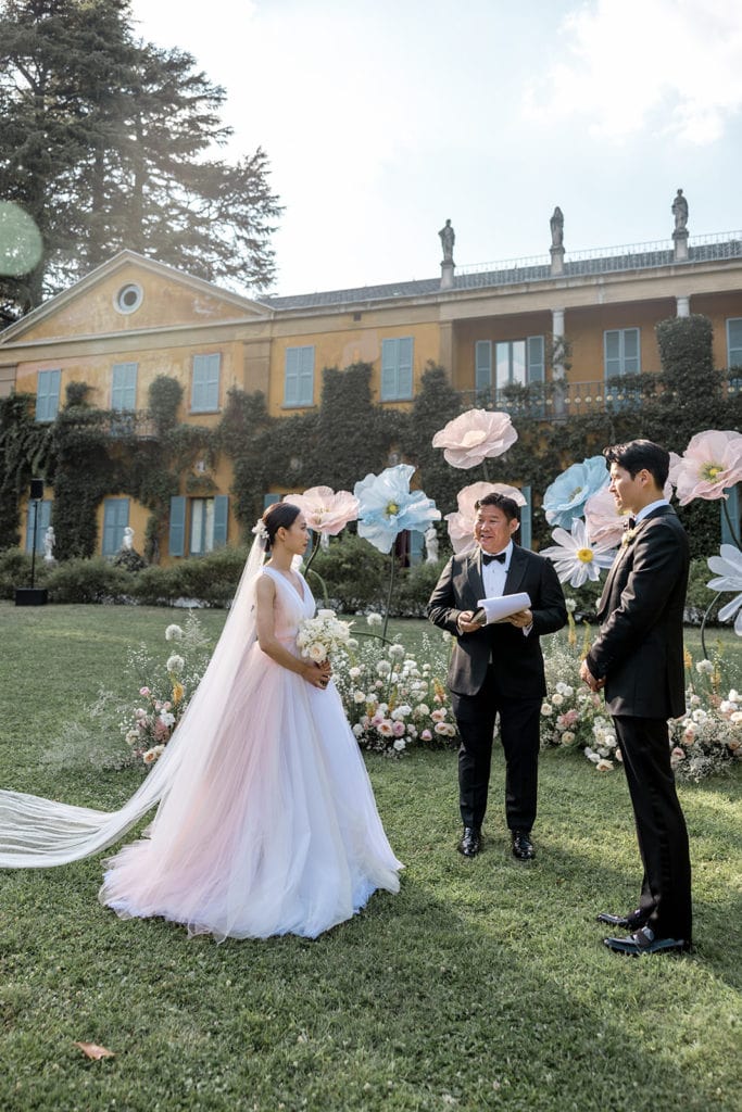 Bride and groom stand in front of whimsical flower statues during Villa Gastel wedding ceremony