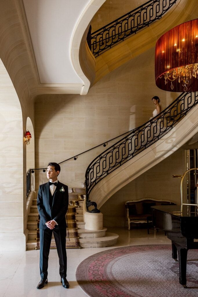 Groom waits for bride for first look as bride walks down staircase before French chateau wedding
