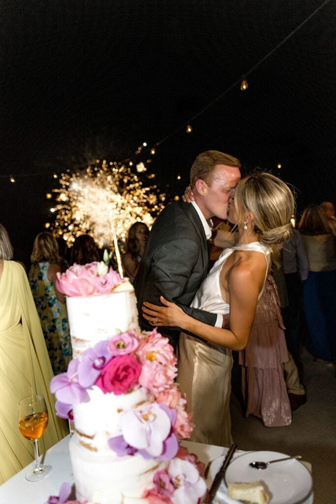 Bride and groom kiss after cutting cake with fireworks in background