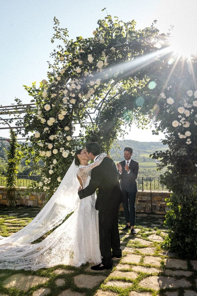 Tuscany wedding ceremony with bride and groom kissing under floral arch