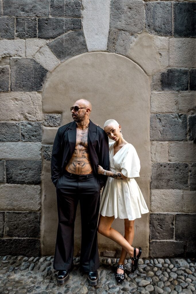 Man shows off tattoos with open suit jacket while woman stands next to him in Lake Como